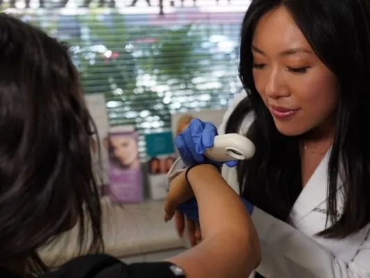 A clinician in a white coat examines a patient’s forearm with a handheld dermatoscope
